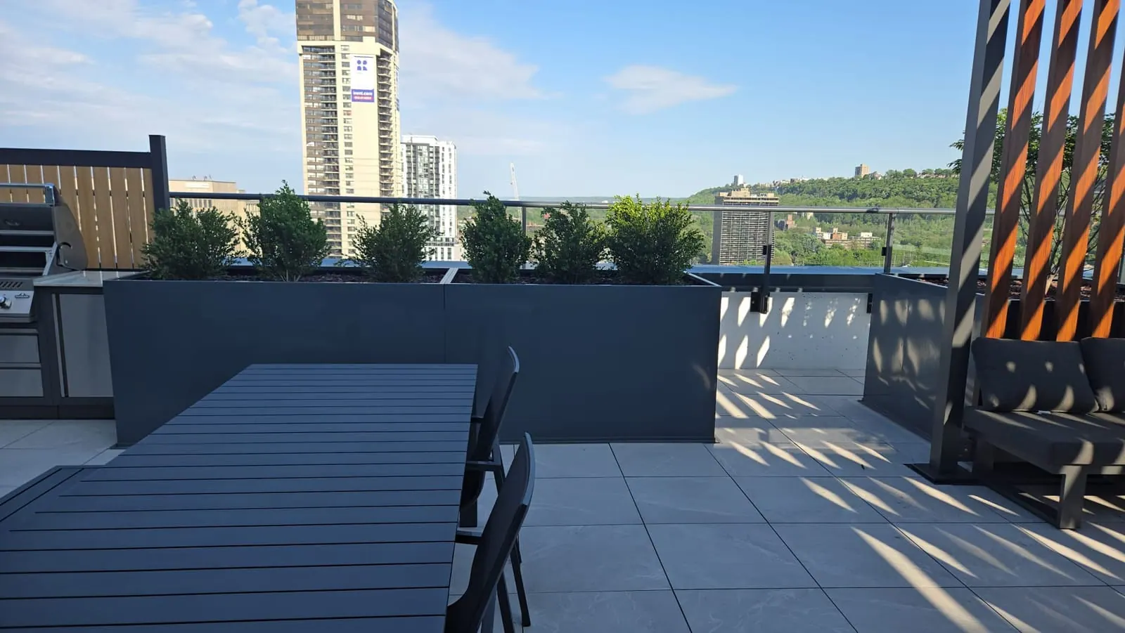 Sunny rooftop terrace with an outdoor dining table, green planters and a grill station beyond.