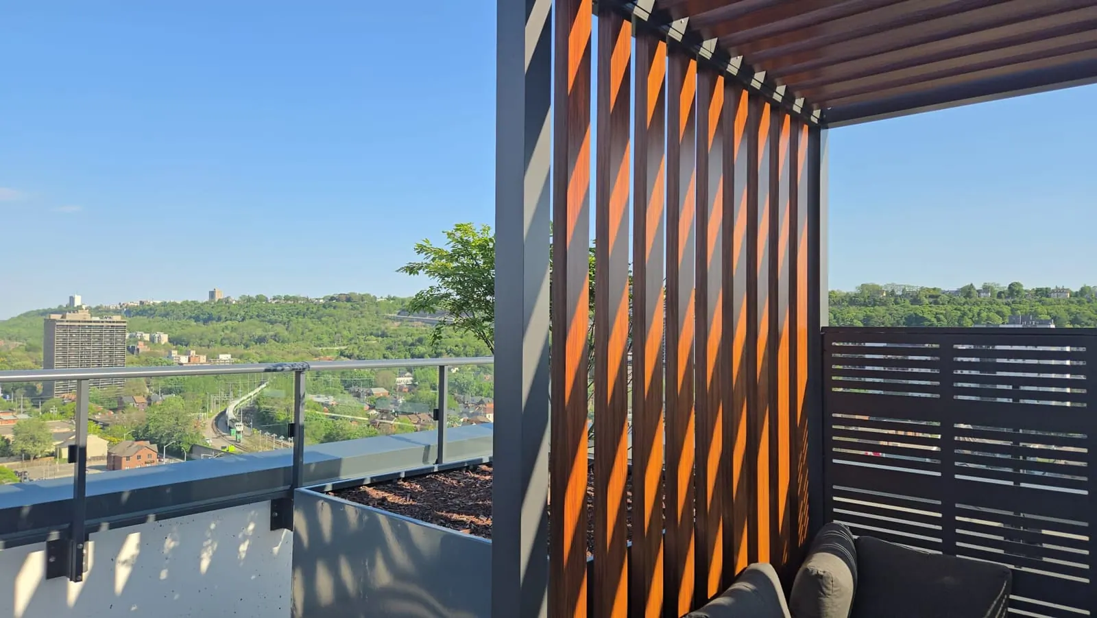 Warm wood-slat pergola screen catching sunlight above a panoramic view of a green river valley and downtown.