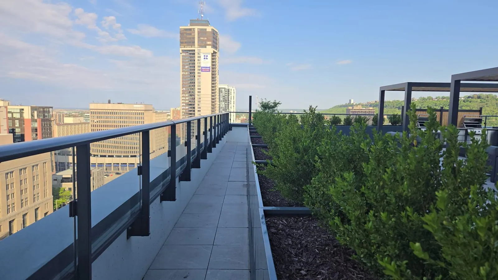 Rooftop walkway between a glass railing and boxwood hedges, leading toward a downtown tower and green hillside.